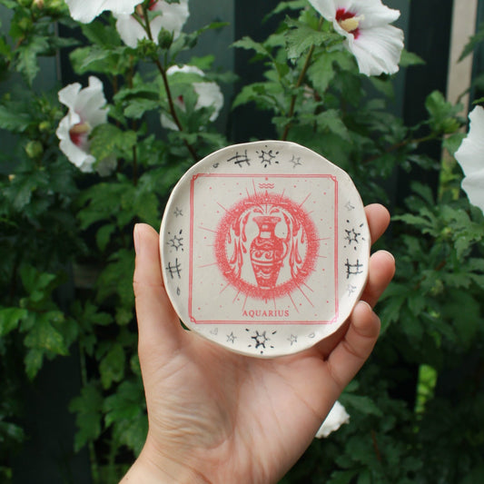 small round jewelry dish decorated with a red square aquarius vase design and surrounded by black pencil doodles. held in hand against a dark foliage background.