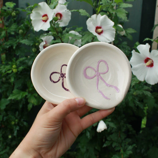 hand thrown off white spoon rests decorated with a purple bow in the centre. held in hand against a dark green foliage background, with white and pink hibiscus flowers peeking out. 