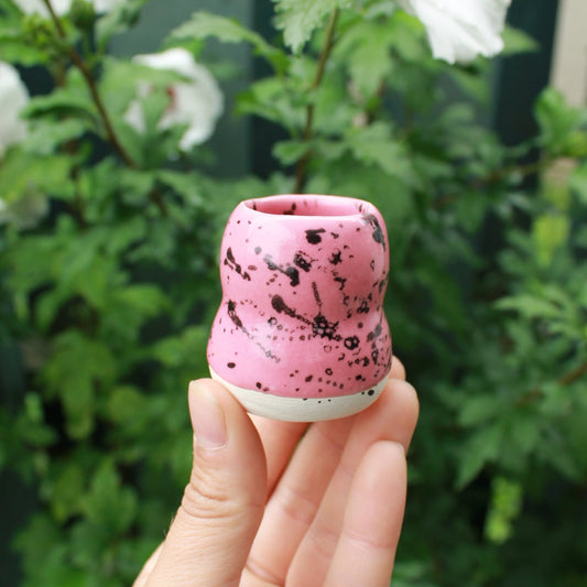 micro bubble bud vase decorated with pink glaze and black splatter. held in hand against a dark foliage background with hibiscus flowers. 