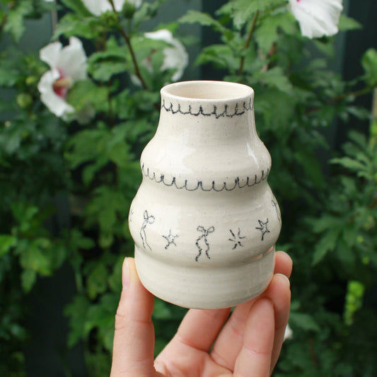 handthrown bubble vase with long neck, decorated with bows, starbursts and scalloped lines. held in hand against a dark foliage background and hibiscus flowers.