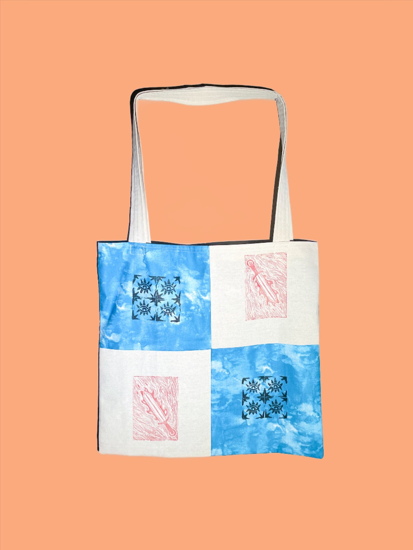 canvas tote bag sits against a peach orange backdrop. the bag is quilted in four simple squares with black and red block printed designs on each square. the canvas squares have a red sword print, and the tie dye blue squares have a quilted eye block print