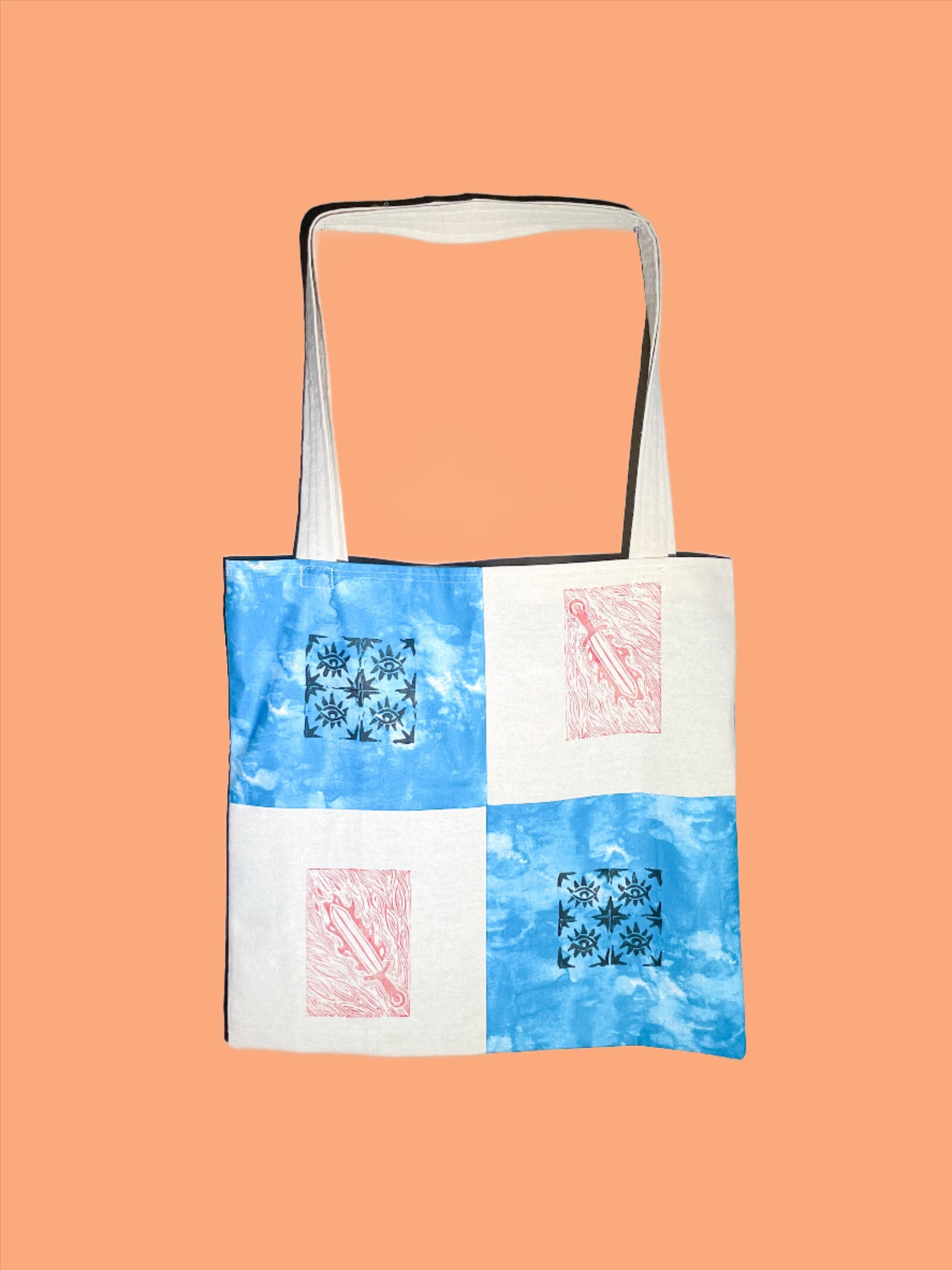 canvas tote bag sits against a peach orange backdrop. the bag is quilted in four simple squares with black and red block printed designs on each square. the canvas squares have a red sword print, and the tie dye blue squares have a quilted eye block print