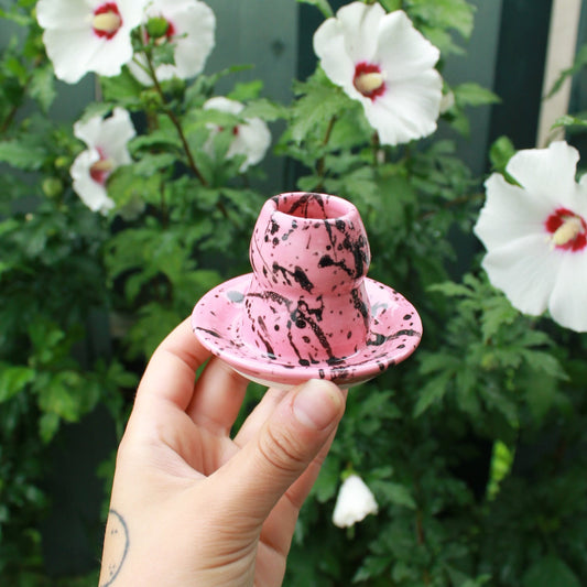 small, curvy, pink candle holder with black splattered glaze. held in hand against a deep green foliage background with white and pink hibiscus flowers peeking out. 
