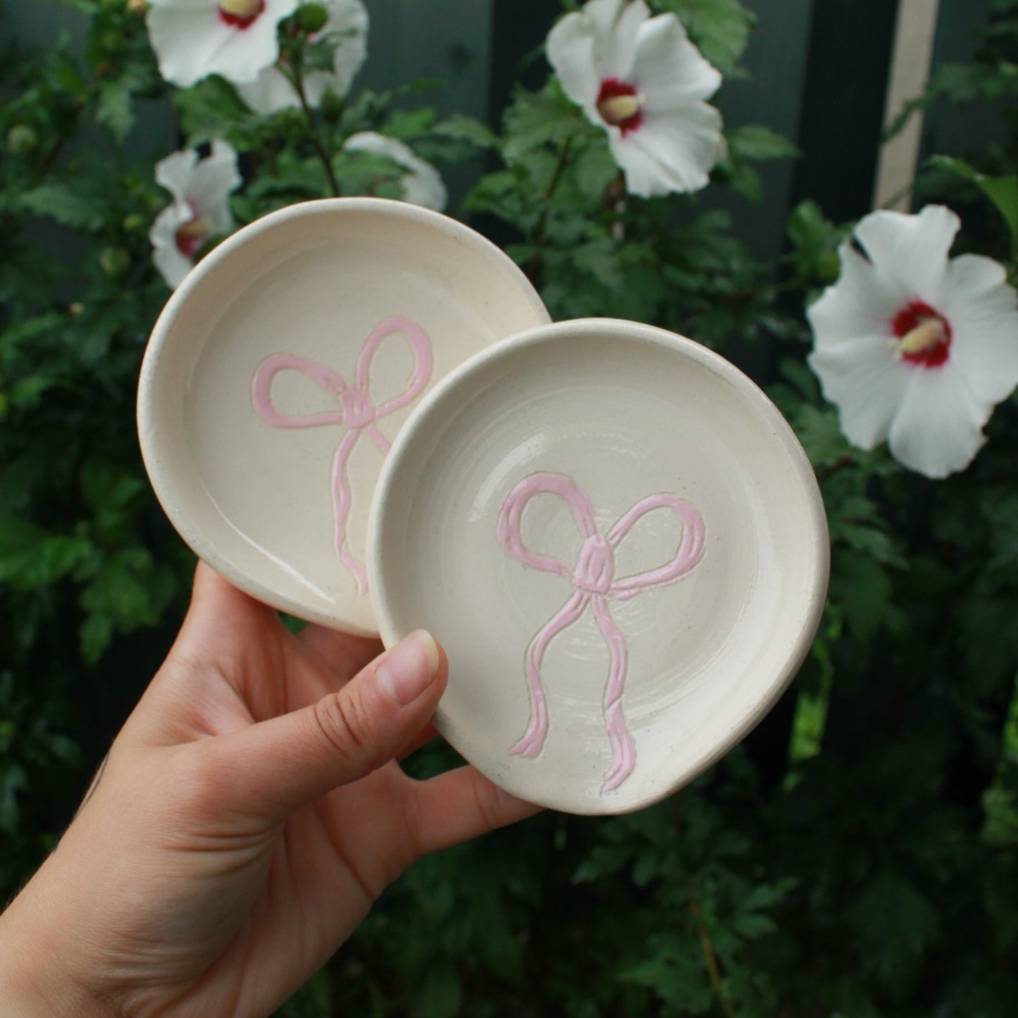 spoon rests decorated with baby pink bows. held in hand against a dark foliage background with white and pink hibiscus flowers. 