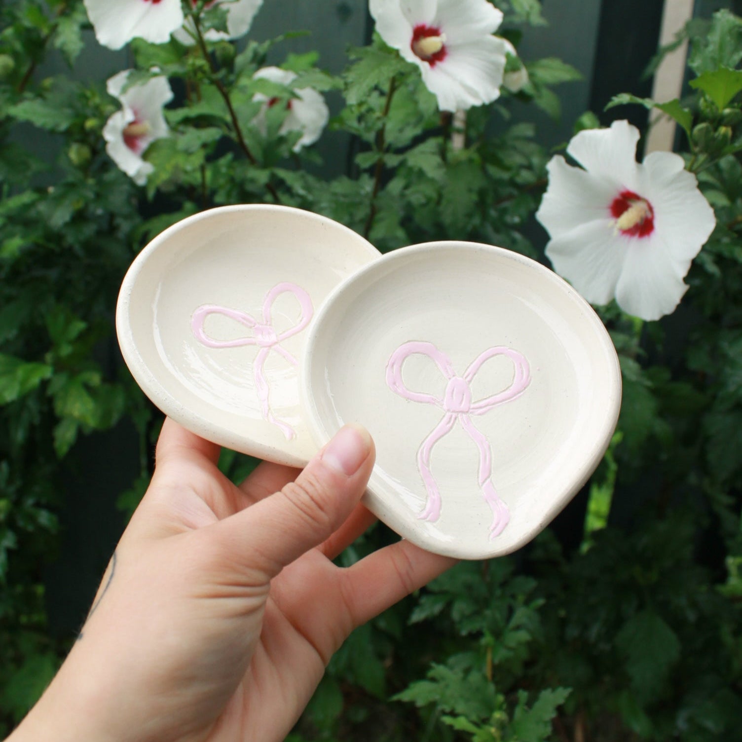 spoon rests decorated with baby pink bows. slightly angled to see the texture of the carved bows. held in hand against a dark foliage background with white and pink hibiscus flowers. 