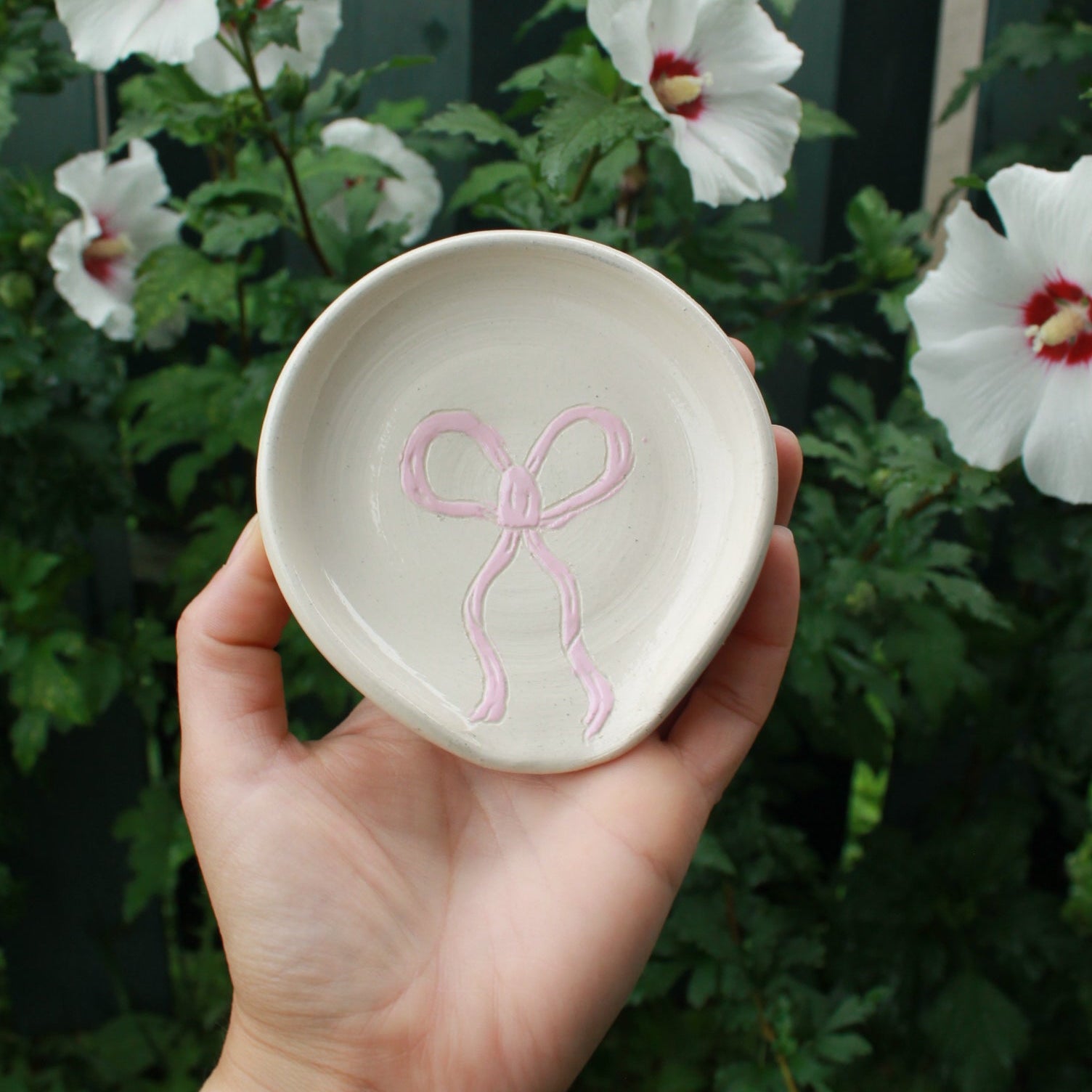 one spoon rest decorated with a baby pink bow. held in hand against a dark foliage background with white and pink hibiscus flowers. 