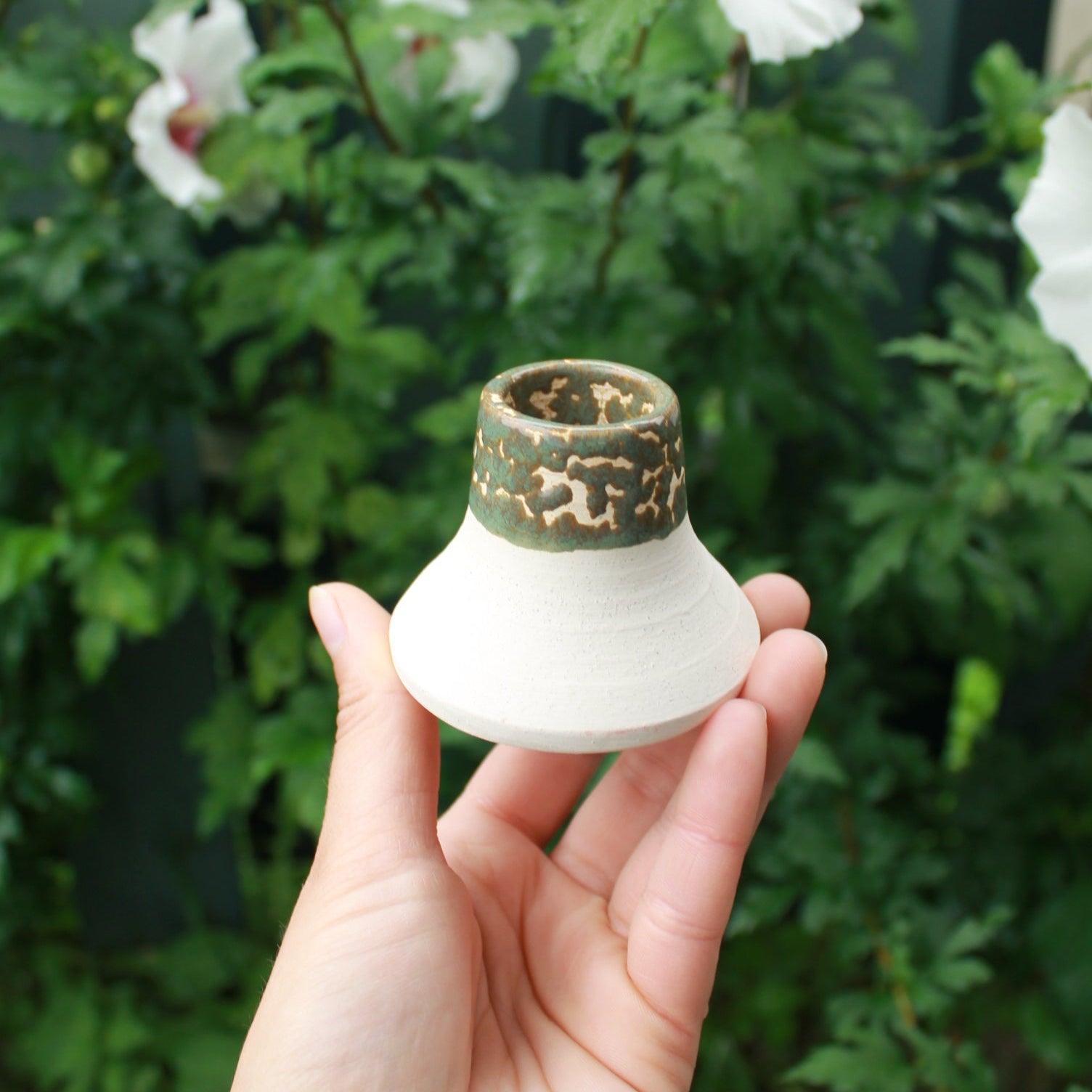 white clay candle holder with crawling sage green glaze. held in hand against a dark foliage background with hibiscus flowers.