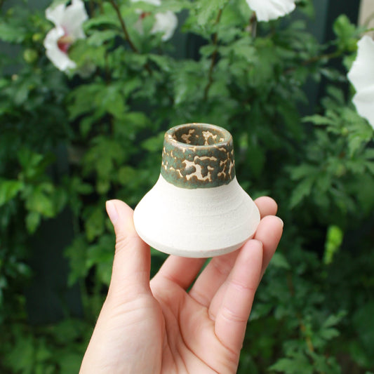 white clay candle holder with crawling sage green glaze. held in hand against a dark foliage background with hibiscus flowers.