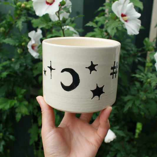 large kitchen utensil jug handthrown from white clay and decorated with black glaze celestial designs. held in hand against a dark foliage background with hibiscus flowers. 