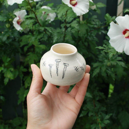tiny vase decorated with a small amount of hatching and messy drawn nails. held in hand against a dark foliage background with white and pink hibiscus flowers peeking out. 