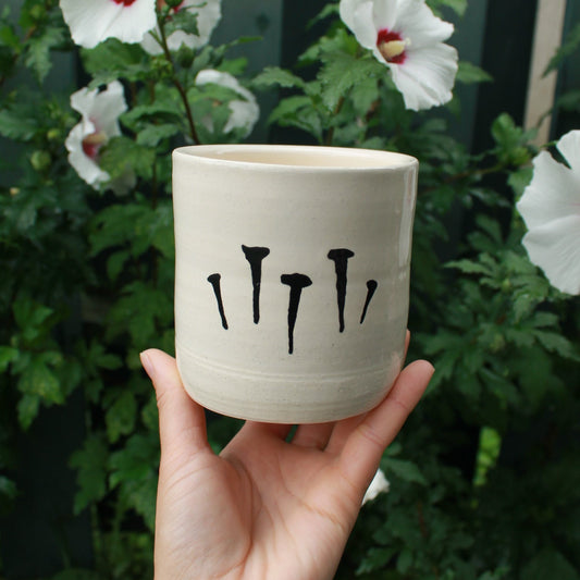 medium sized hand thrown kitchen utensil cup decorated with 5 black glazed nails. held in hand against a dark foliage background with white hibiscus flowers. 