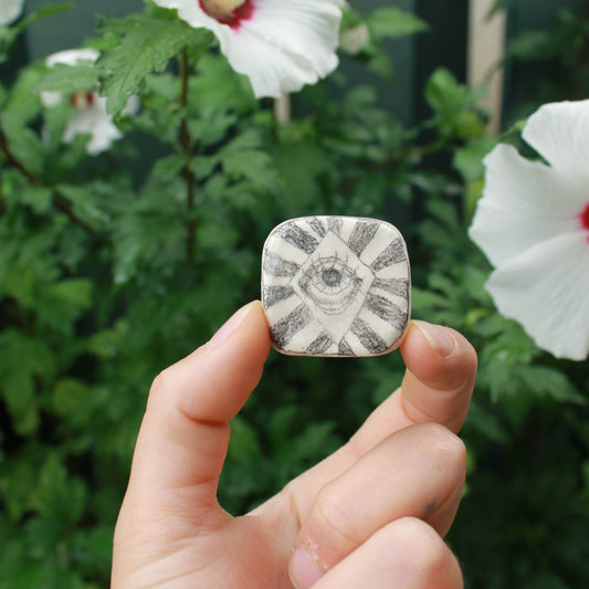 tiny square bud vase decorated with an eye and stripes, held in hand against a dark foliage background and hibiscus flowers.