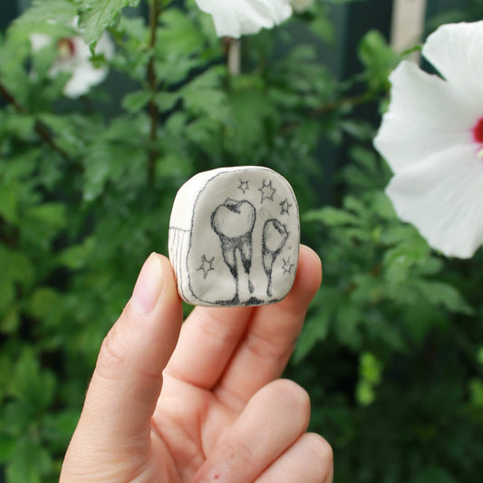 miniature square bud vase decorated with underglaze pencil drawn teeth and starbursts. held in hand against a background of dark foliage and hibiscus flowers. 