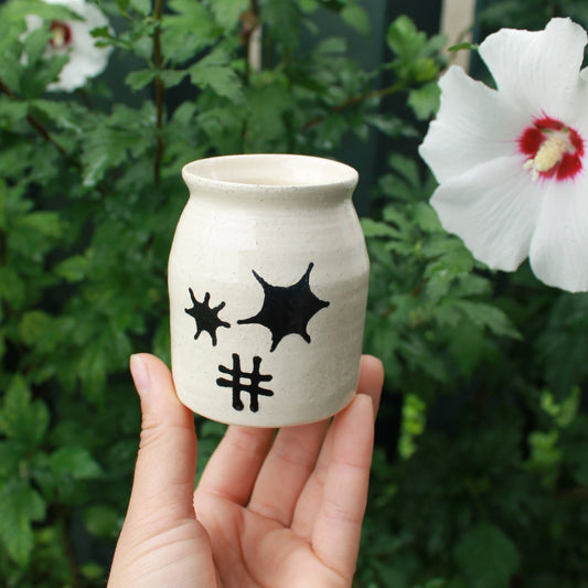 small white jar bud vase decorated with black starburst and hatch designs in the centre. held in hand against a dark foliage backdrop with hibiscus flowers.