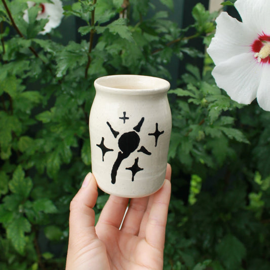 little jar shaped bud vase hand thrown from white clay. decorated with a drippy sun detail and starbursts in black glaze. held in hand against a dark foliage background with white hibiscus flowers. 