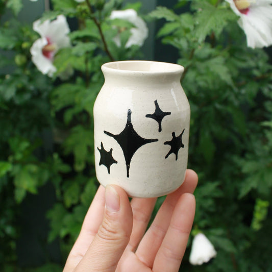 small jar vase made from white clay, decorated with black glaze stars in different shapes and sizes. held in hand against a dark foliage backdrop with white hibiscus flowers.