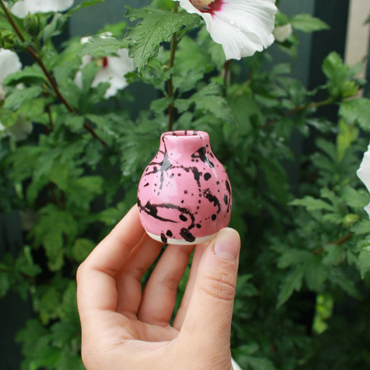 tiny pink bud vase with a shoulder and small neck, decorated with pink glaze and black splatter. held in hand against a dark foliage background with hibiscus flowers. 