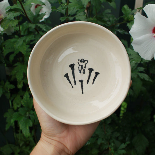 Hand holding a ceramic bowl with black glaze painted tooth and nails against a green leafy background