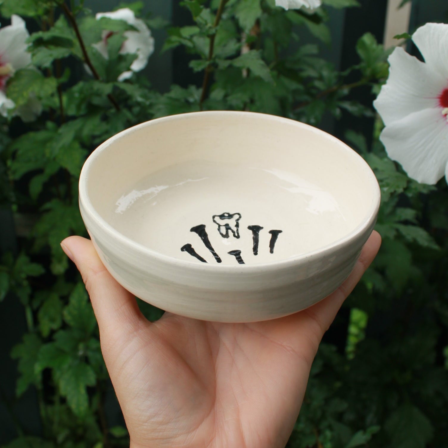 Hand holding a ceramic bowl on an angle with black glaze painted tooth and nails against a green leafy background