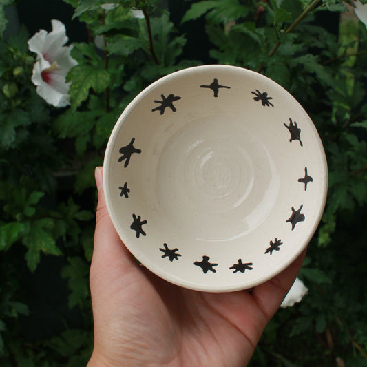 small white bowl with black stars decorating the inside rim. held in hand against a dark foliage background with hibiscus flowers. 